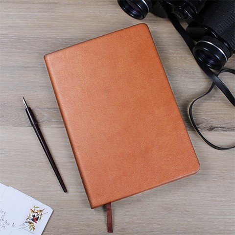 A closed "Custom Medical Journal: Personalized Notebook for Doctors & Nurses - Leather" lies on a wooden surface. To the left of the notebook is a black fountain pen, and a piece of paper with a colorful drawing is partially visible. At the top right are black binoculars with a neck strap. This unique gift makes for thoughtful and stylish writing moments.